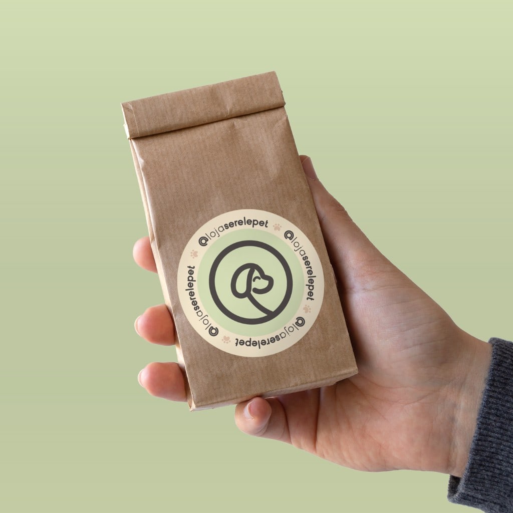 A hand holds a small folded kraft pouch against a sage green background. A round sticker shows a minimal dog profile, cream and green rings, and @lojaserelepet repeated around the edge with small paw prints.